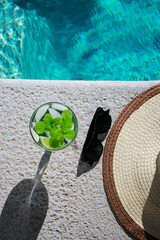 cold mojito cocktail with limes at pool with sunscreen, sunglasses and straw hat