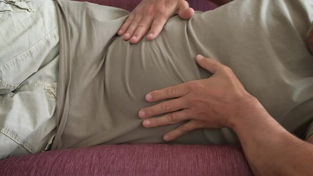 Suffering from abdominal pain in loneliness. A man lay on the sofa and rubbing his belly because of pain in the room.