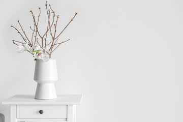 Vase with tree branches and flowers on table near light wall