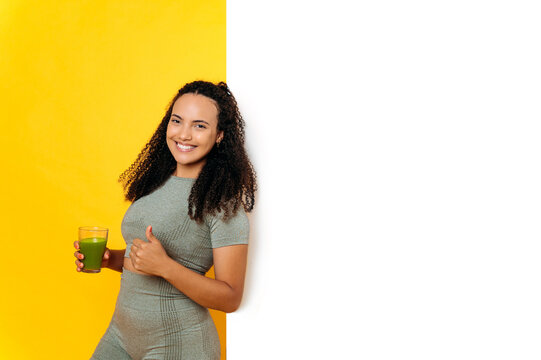 Positive brazilian curly sporty young woman, in sportswear, holds a glass of fresh healthy green smoothie, shows thumb up, stands on isolated yellow background near a white space for a presentation