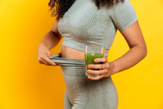 The concept of diet, weight loss, healthy eating, detox. Woman in sportswear, holds a glass of fresh healthy green smoothie in hand, touching her leggings, indicating weight loss, isolated background