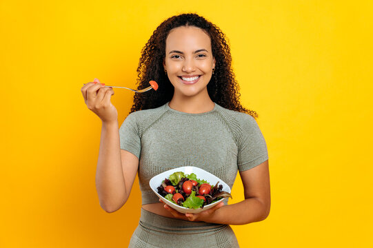 Healthy eating concept. Happy, pretty brazilian or hispanic curly haired sporty woman, in sports outfit, holds fresh salad with tomatoes, standing on isolated yellow background, eats vegetables, smile