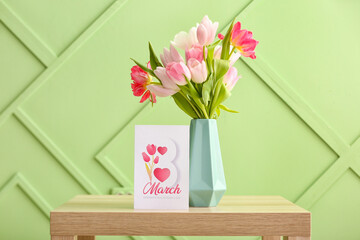 Vase with tulips and greeting card for Women's Day on table near green wall