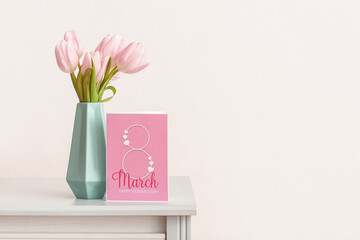 Vase with tulips and greeting card for Women's Day on shelf near light wall