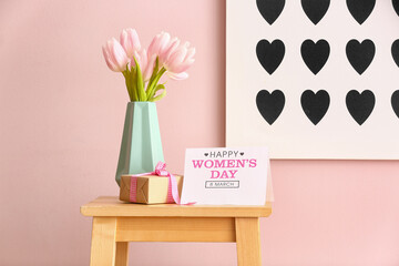 Vase with tulips, gift and greeting card for Women's Day on table near pink wall