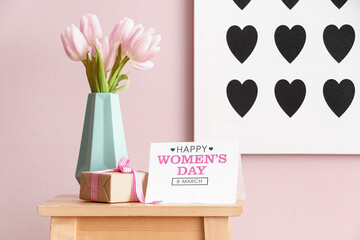 Vase with tulips, gift and greeting card for Women's Day on table near pink wall