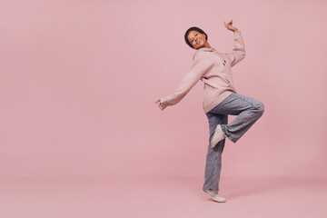 Fototapeta na wymiar Young smiling black woman dancing and jumping over pink pastel background. Cute happy girl wearing jeans and hoodie posing at camera