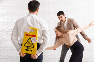 Young man with caution sign playing a prank on his colleagues against white brick background. April...