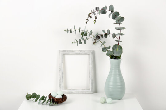 Blank frame, vase with eucalyptus branches and Easter eggs on white background