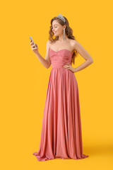 Young woman in tiara and prom dress using mobile phone on yellow background
