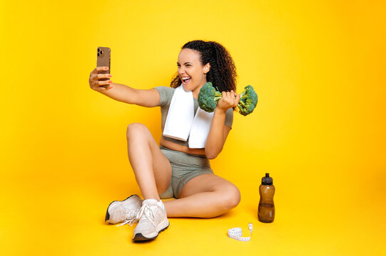 Full length happy funny brazilian or hispanic curly woman in sports outfit, takes selfie on smartphone with broccoli dumbbell in hand while sitting on isolated orange background, smiling, having fun