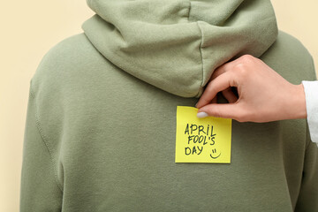 Woman sticking paper with text APRIL FOOL'S DAY on friend's back against color background, closeup