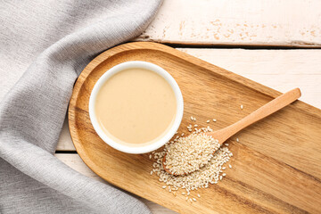 Board with bowl of tasty tahini and sesame seeds on light wooden background, closeup