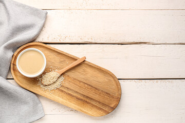 Board with bowl of tasty tahini and sesame seeds on light wooden background