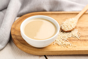 Board with bowl of tasty tahini and sesame seeds on light wooden table, closeup