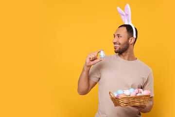 Happy African American man in bunny ears headband holding wicker tray with Easter eggs on orange...