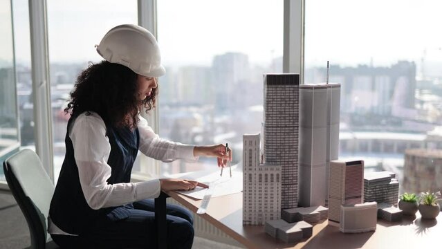 Curly female engineer working on building complex prototype project of residential or business district, measuring blueprint city model using compass at office with panoramic windows
