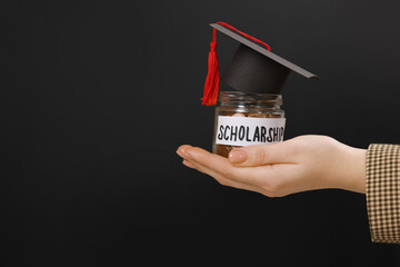 Woman holding glass jar of coins and graduation cap against black background, closeup with space...