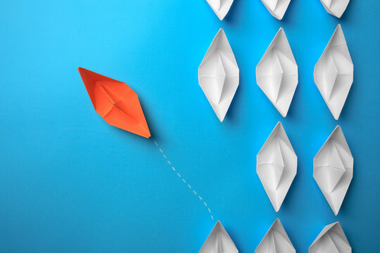 Orange paper boat floating away from others on light blue background, flat lay. Uniqueness concept