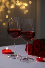 Glasses of red wine, rose flowers and burning candles on white table against blurred lights. Romantic atmosphere