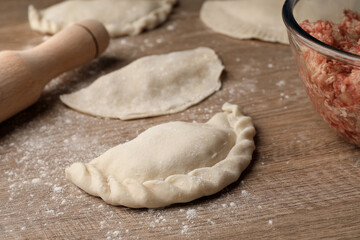Uncooked chebureki, minced meat and rolling pin on wooden table, closeup