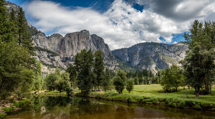 Fototapeta na wymiar Looking across the smooth flat Merced river towards Yosemite Falls with fluffy white clouds and blue sky