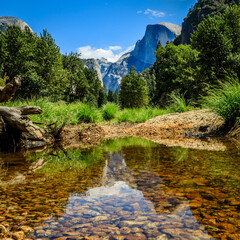 View of half dome looking across the Merced river with smooth water and brown rocks and a bright blue sky