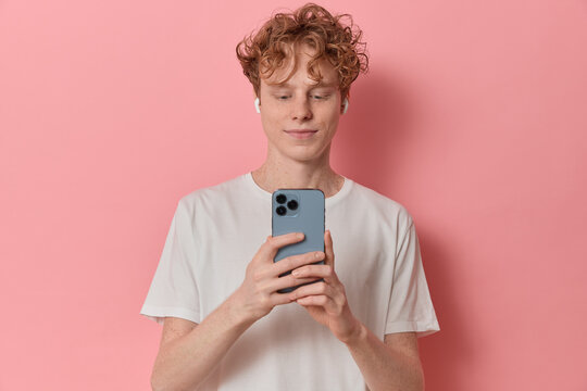 Handsome curly haired young man smiles happily uses mobile phone application taking pictures on a smartphone listens to music with headphones wears casual t-shirt isolated over pink background.