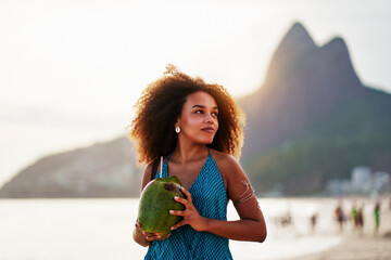 waist up portrait cheerful young brazilian afro hairstyle woman walking on the beach holding a coconut water in Ipanema 	 - obrazy, fototapety, plakaty