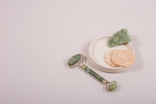 Rodillo de cuarzo de jade y guasha, masajeadores de rostro, en fondo neutro sobre una base org&aacute;nica, y esponjas naturales faciales con espacio para textos