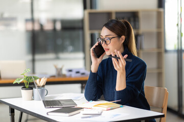 Cheerful business Asian woman freelancer making telephone call share good news about project working in office workplace, business finance concept.	
