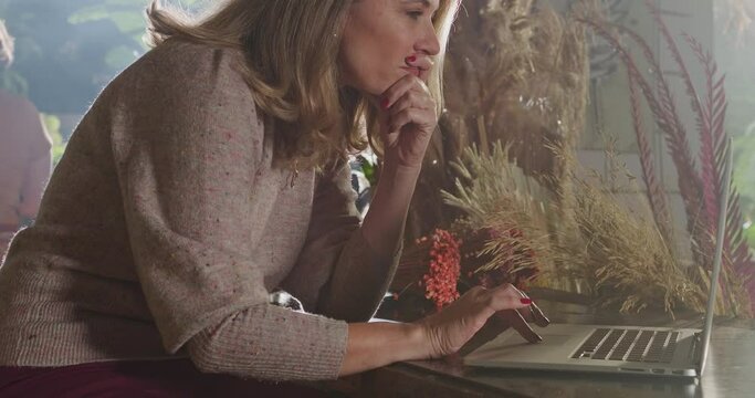Concentrated middle age small business owner reading content online in front of laptop computer taking online orders for delivery at flower shop