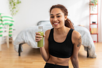 Smiling young woman drinking green fresh detox juice after workout routine at home