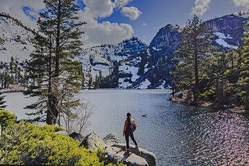 Woman standing near small peaceful lake with snowy mountains on the background - obrazy, fototapety, plakaty
