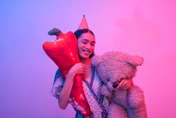 A blissful trans woman wearing a party hat carrying a large teddy bear and chili stuffed toy. Party...