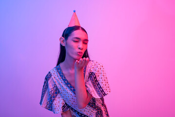 A trans woman closes her eyes while blowing a kiss to the camera. Lit with blue and pink neon colors.