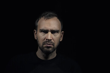 A serious man with grey hair and a small beard looks sullenly into the camera. A portrait of a Caucasian man with frowning eyebrows. A dramatic portrait on a dark background.