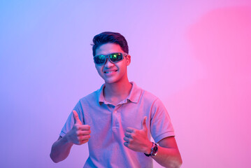 A young asian man wearing shades making two thumbs up sign. Lit with blue and pink neon colors.