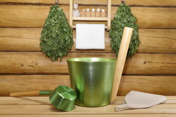 On a wooden bench in sauna there are metal vat, ladle and hat and all these against the background of log wall with birch brooms hanging on it near shelf with bottles of essential oils standing on it.