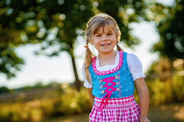Cute little kid girl in traditional Bavarian costume in wheat field. Happy child with hay bale during Oktoberfest in Munich. Preschool girl play at hay bales during summer harvest time in Germany.
