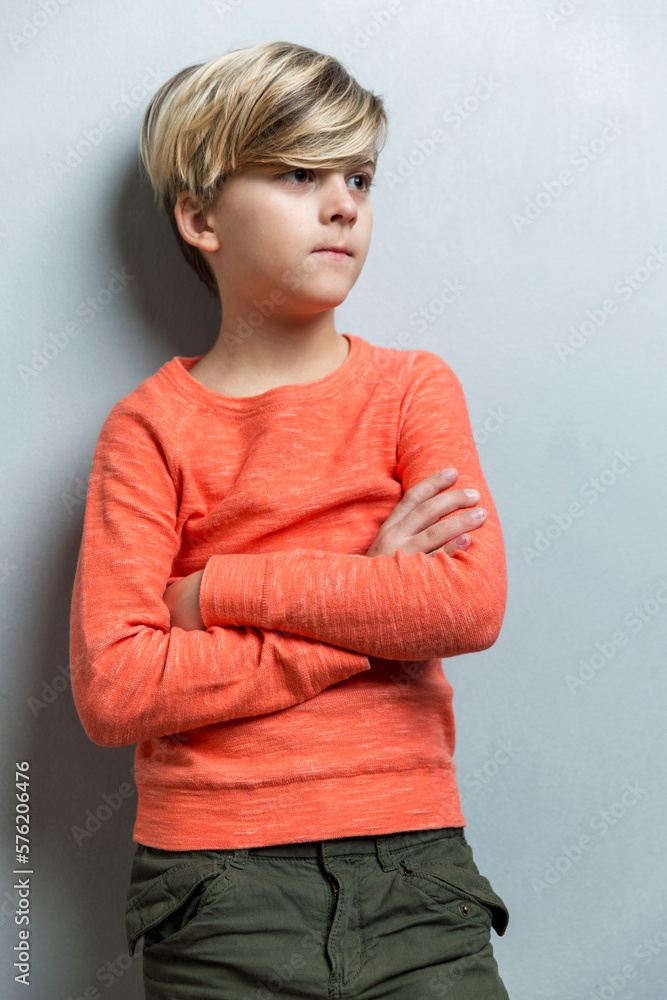 Wall mural A serious boy of 9-10 years old stands near a gray wall. The guy in the orange sweater crossed his arms over his chest. Vertical.