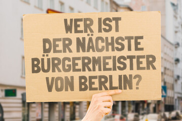 The phrase " Who's the next mayor of Berlin? " on a banner in men's hand with blurred background. Election. City management. Politics. Urban. Voter. Candidate