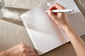 Women's hand writing in blank notebook sheet with sunlight shadows, home office lifestyle...