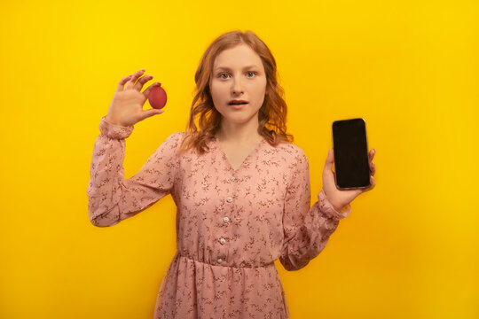 Surprised shocked young woman in pink dress holding red painted dyed easter egg and showing empty mobile phone black screen mock up isolated on yellow background.

Easter celebration, spring concept.