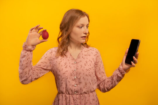 Surprised astonished shocked girl in pink dress holds red painted dyed easter egg and looks at empty mobile phone black screen mockup isolated on yellow background.

Easter day or spring concept.