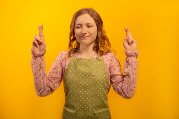 Young woman in green polka dot kitchen apron hold fingers crossed for good luck, plead God,...