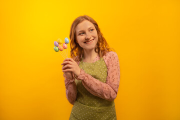 Happy smiling cheerful housewife girl in green polka dot kitchen apron holding small multicolored painted dyed easter eggs sticks and looking aside isolated on yellow background.

Easter day concept.