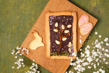 Polish Easter sweet Mazurek made of shortbread dough stuffed with chocolate, decorated with nuts, dried fruits and sugar sprinkles on a wooden board on a green concrete background.