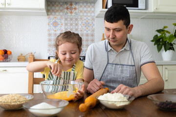 Obraz na płótnie Canvas child helping to father in kitchen. kid cooking food with dad. little girl, man in apron in preparing dough, baking pie, cookies, making biscuit. family together home