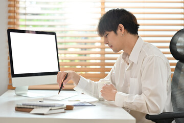 Thoughtful asian male investor sitting at his workplace front of blank computer screen and reading agreement before signing